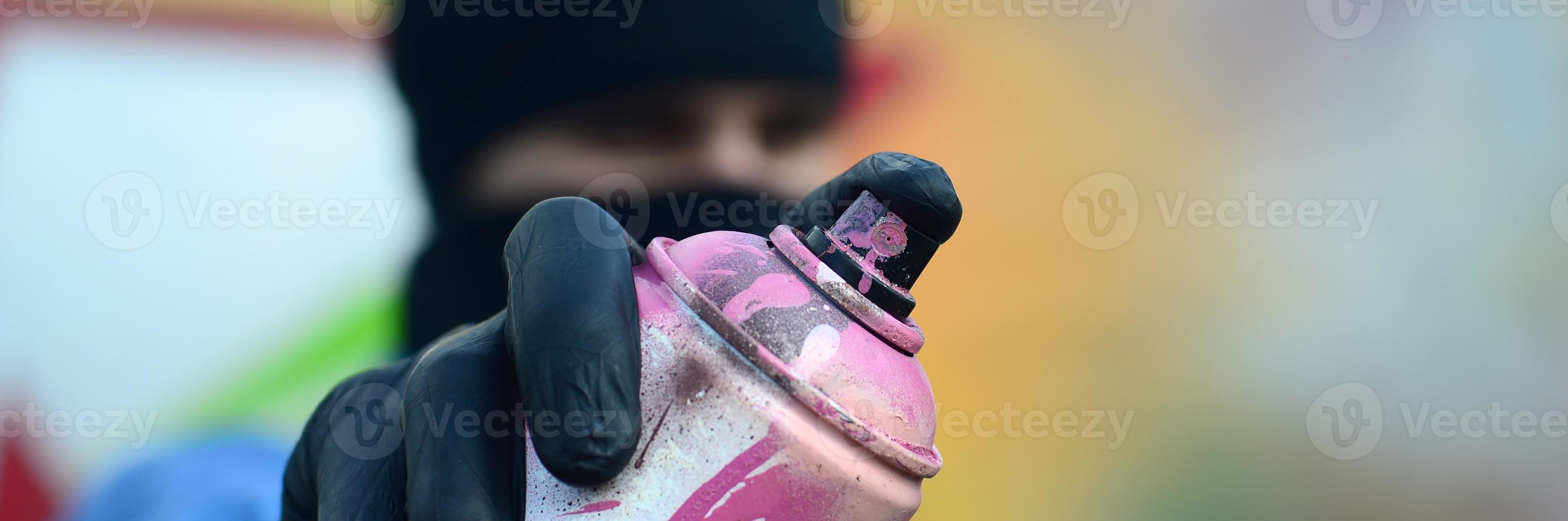 A young graffiti artist in a blue jacket and black mask is holding a can of paint in front of him against a background of colored graffiti drawing. Street art and vandalism concept photo