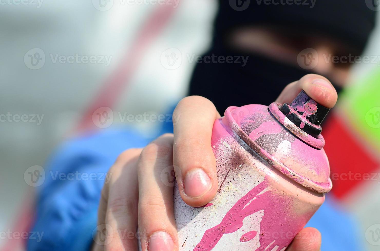 A young graffiti artist in a blue jacket and black mask is holding a can of paint in front of him against a background of colored graffiti drawing. Street art and vandalism concept photo