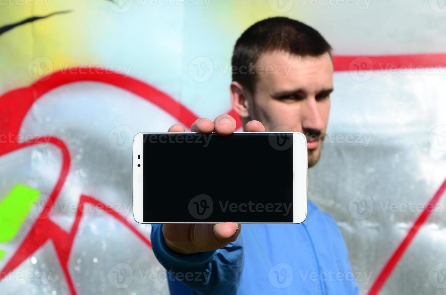 The graffiti artist demonstrates a smartphone with an empty black screen against the background of a colorful painted wall. Street art concept photo