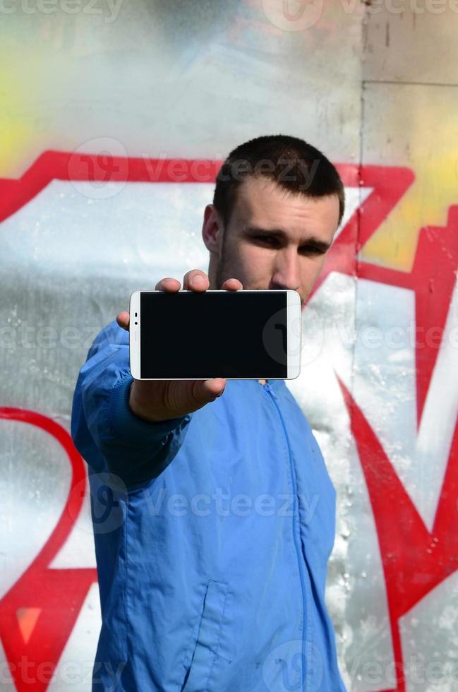 The graffiti artist demonstrates a smartphone with an empty black screen against the background of a colorful painted wall. Street art concept photo