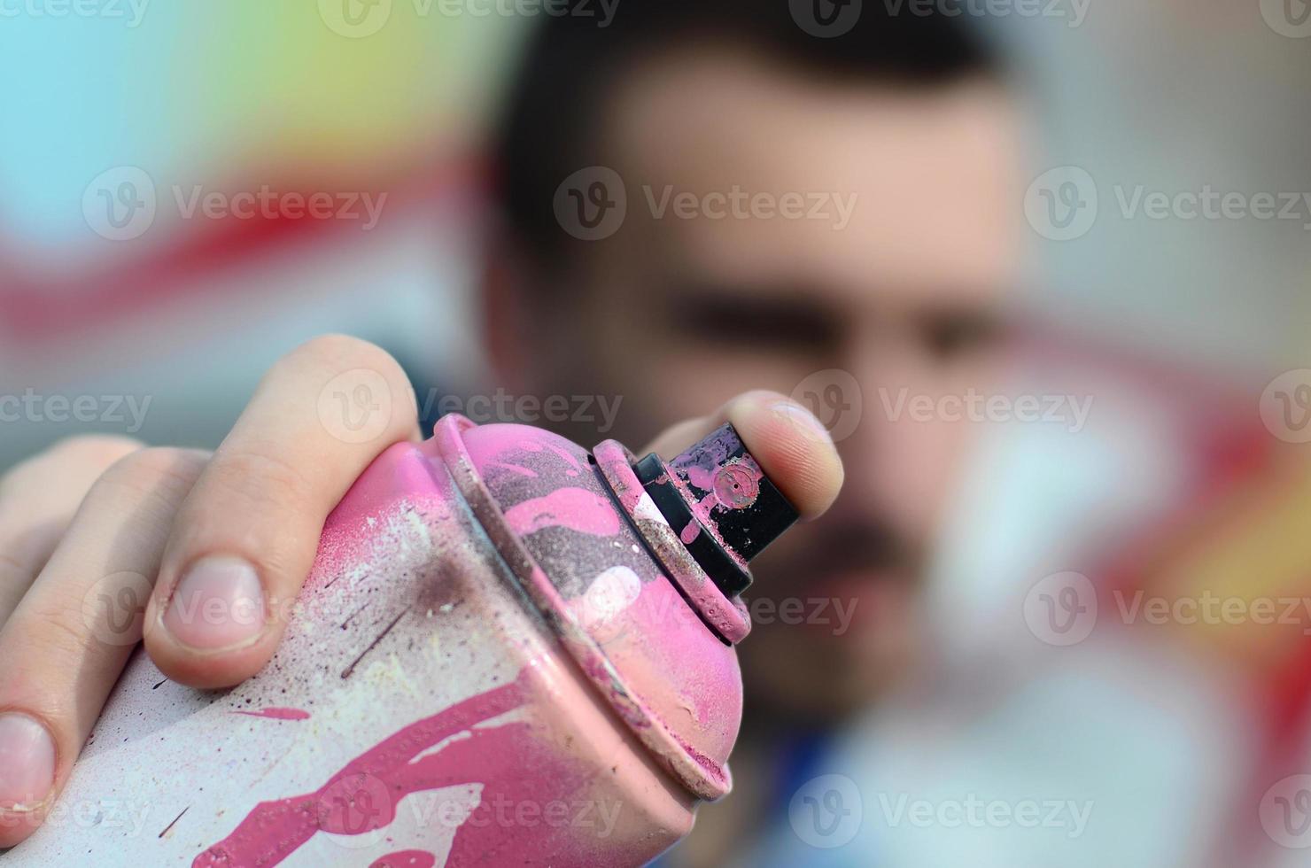 A young graffiti artist in a blue jacket is holding a can of paint in front of him against a background of colored graffiti drawing. Street art and vandalism concept photo