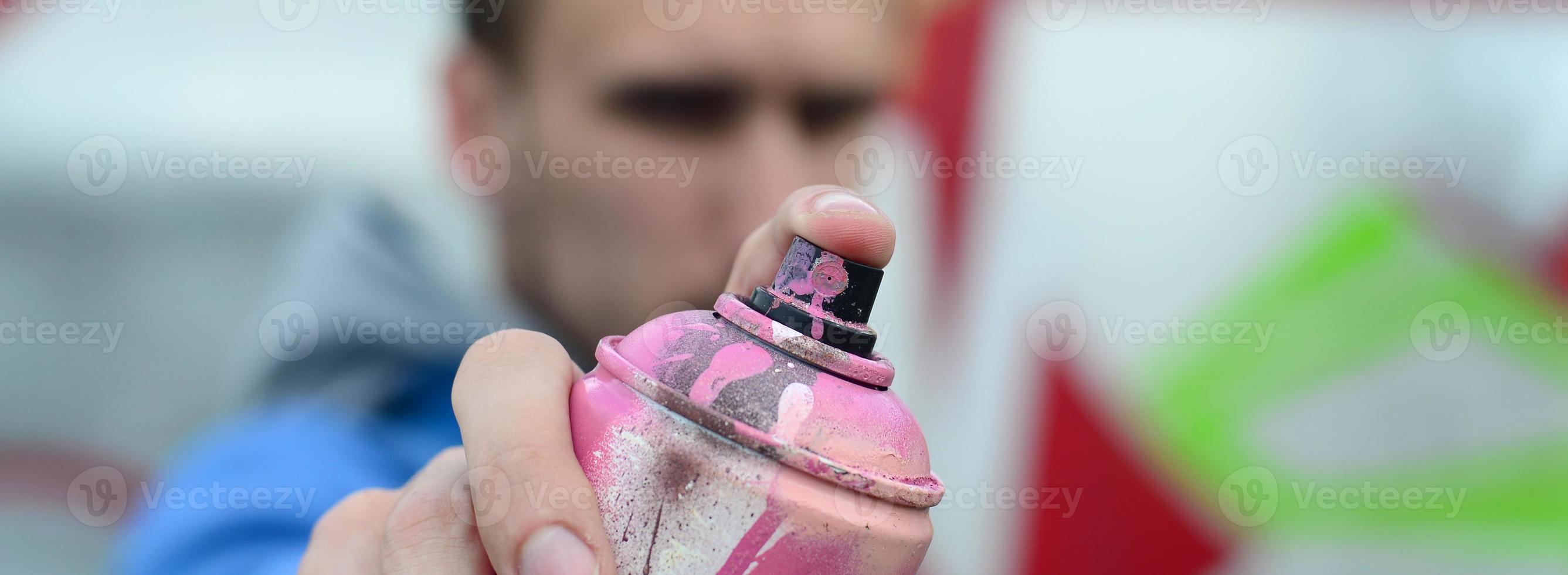 A young graffiti artist in a blue jacket is holding a can of paint in front of him against a background of colored graffiti drawing. Street art and vandalism concept photo