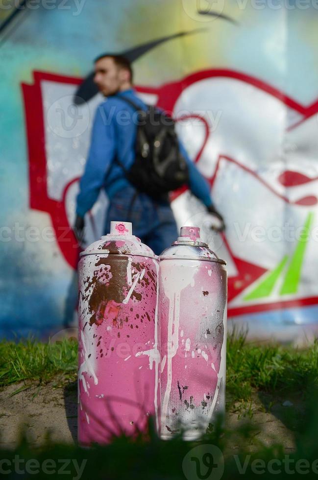 A few used paint cans against the background of the space with the wall on which the young guy draws a large graffiti drawing. Modern art of drawing walls in graffiti photo