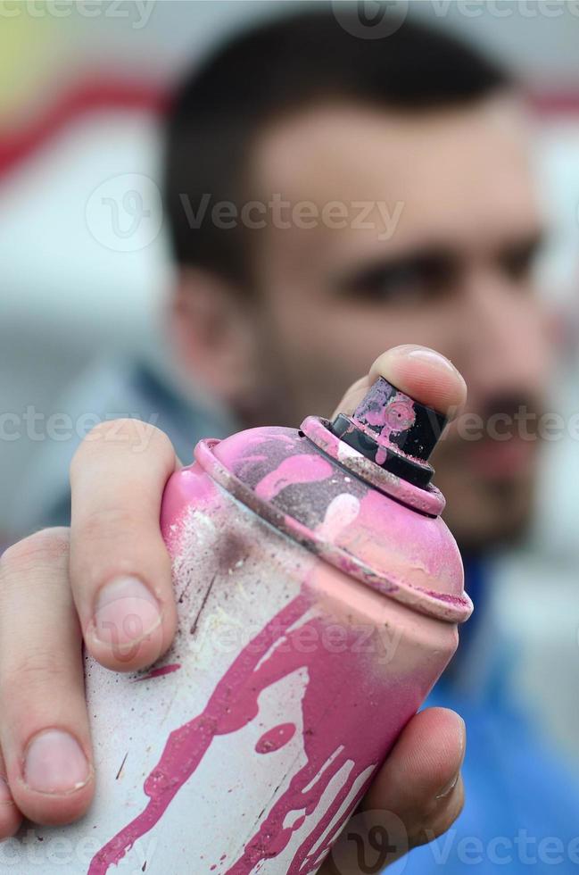 A young graffiti artist in a blue jacket is holding a can of paint in front of him against a background of colored graffiti drawing. Street art and vandalism concept photo