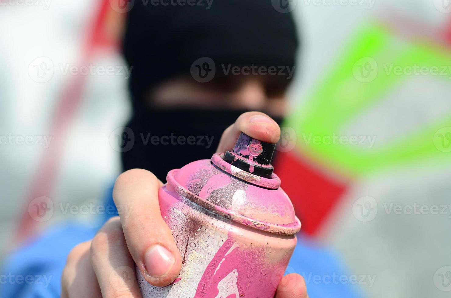 A young graffiti artist in a blue jacket and black mask is holding a can of paint in front of him against a background of colored graffiti drawing. Street art and vandalism concept photo