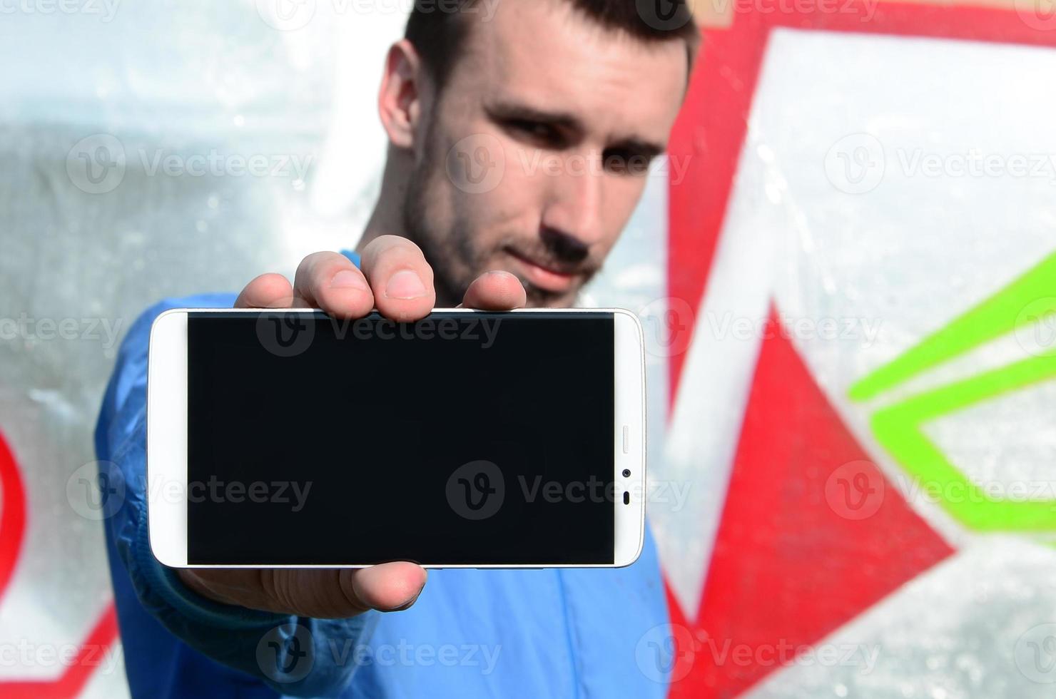 The graffiti artist demonstrates a smartphone with an empty black screen against the background of a colorful painted wall. Street art concept photo