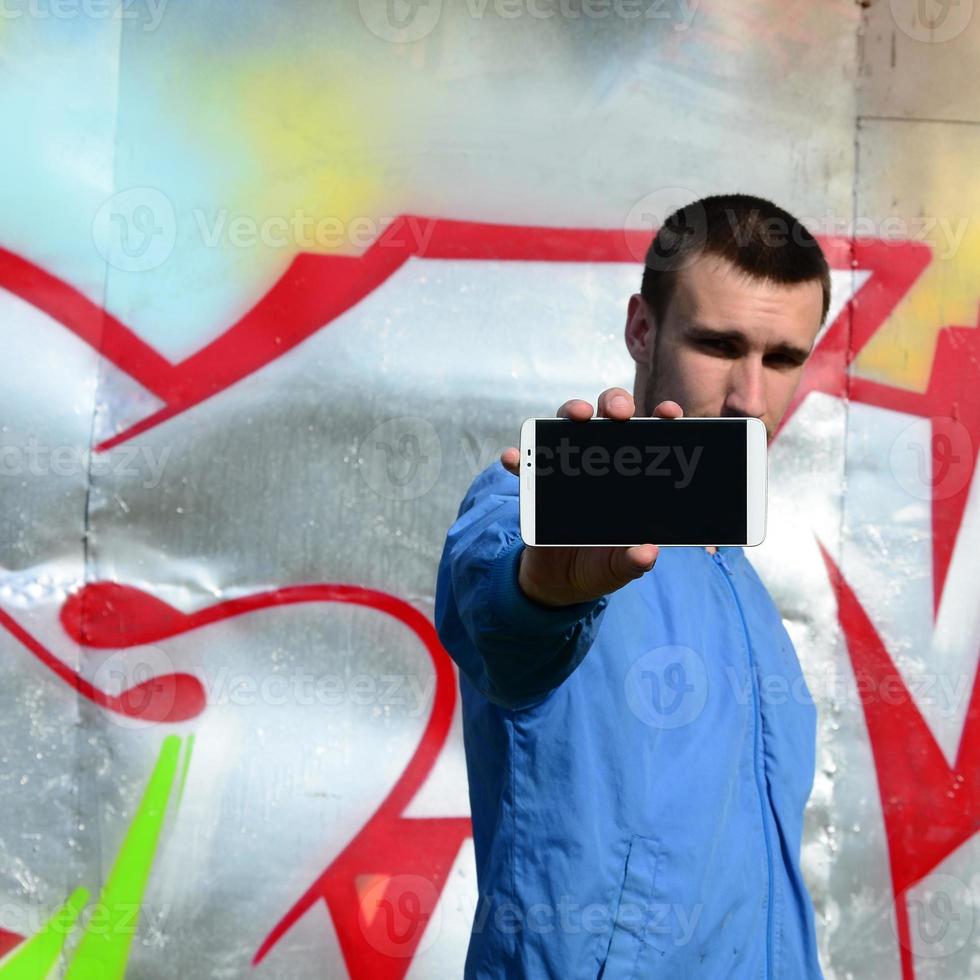 The graffiti artist demonstrates a smartphone with an empty black screen against the background of a colorful painted wall. Street art concept photo