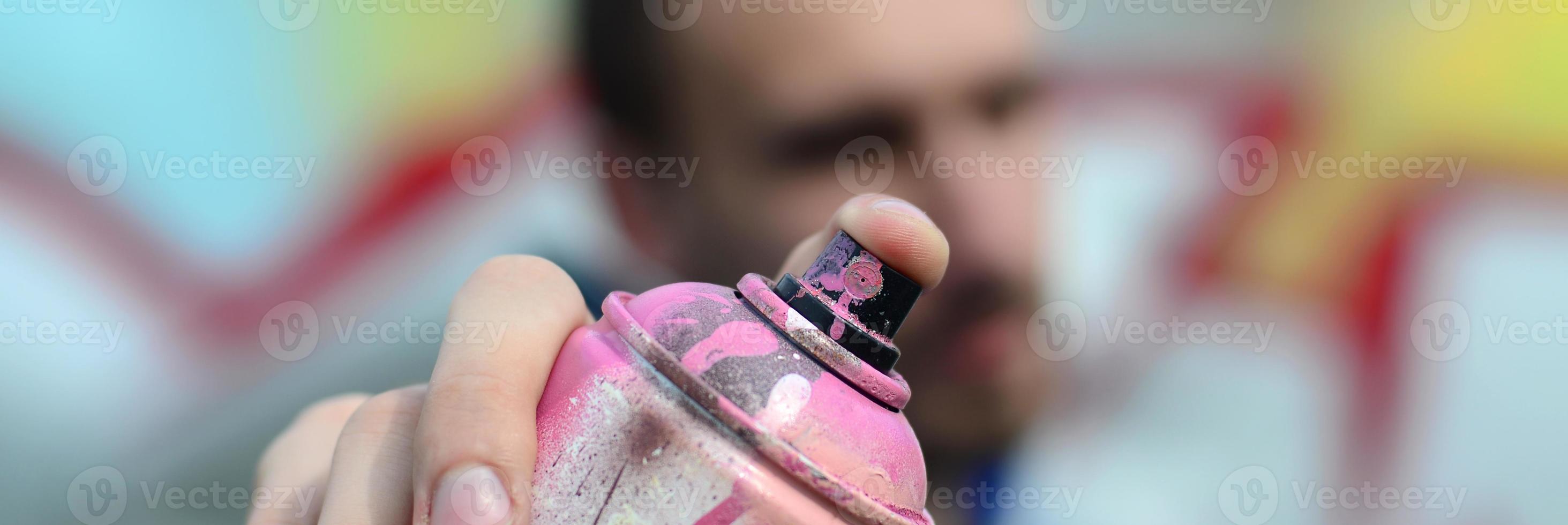 A young graffiti artist in a blue jacket is holding a can of paint in front of him against a background of colored graffiti drawing. Street art and vandalism concept photo