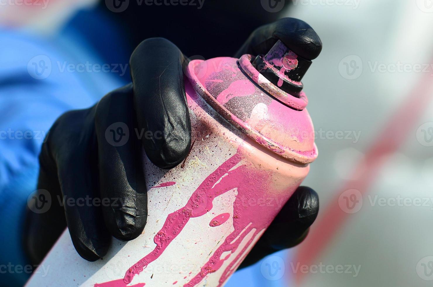 A young graffiti artist in a blue jacket and black mask is holding a can of paint in front of him against a background of colored graffiti drawing. Street art and vandalism concept photo