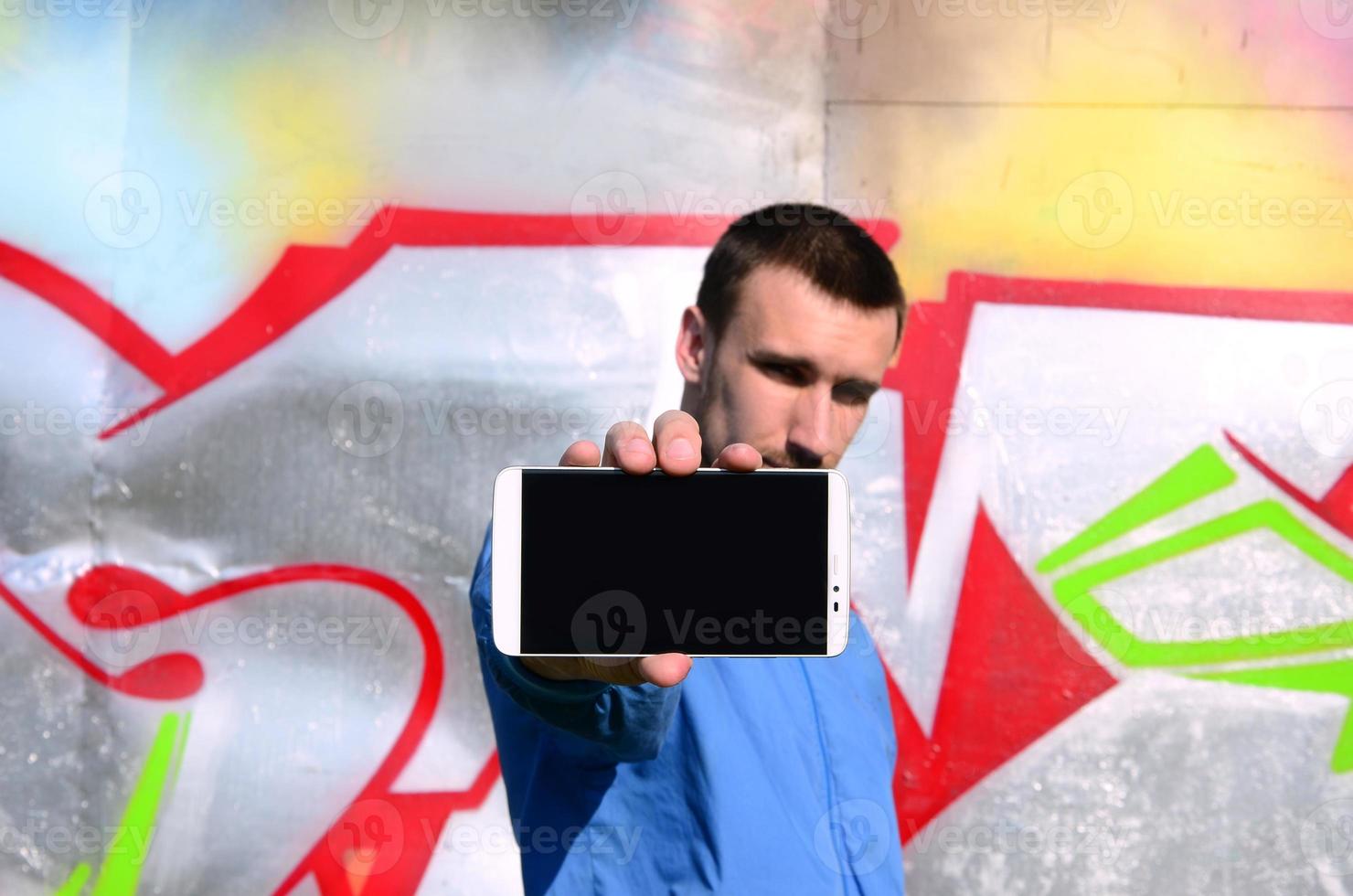 The graffiti artist demonstrates a smartphone with an empty black screen against the background of a colorful painted wall. Street art concept photo