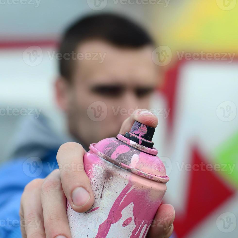 un joven artista de graffiti con una chaqueta azul sostiene una lata de pintura frente a él sobre un fondo de dibujo de graffiti de colores. concepto de arte callejero y vandalismo foto