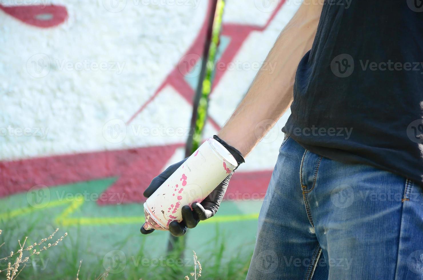 A young hooligan with a spray can stands against a concrete wall with graffiti paintings. Illegal vandalism concept. Street art photo