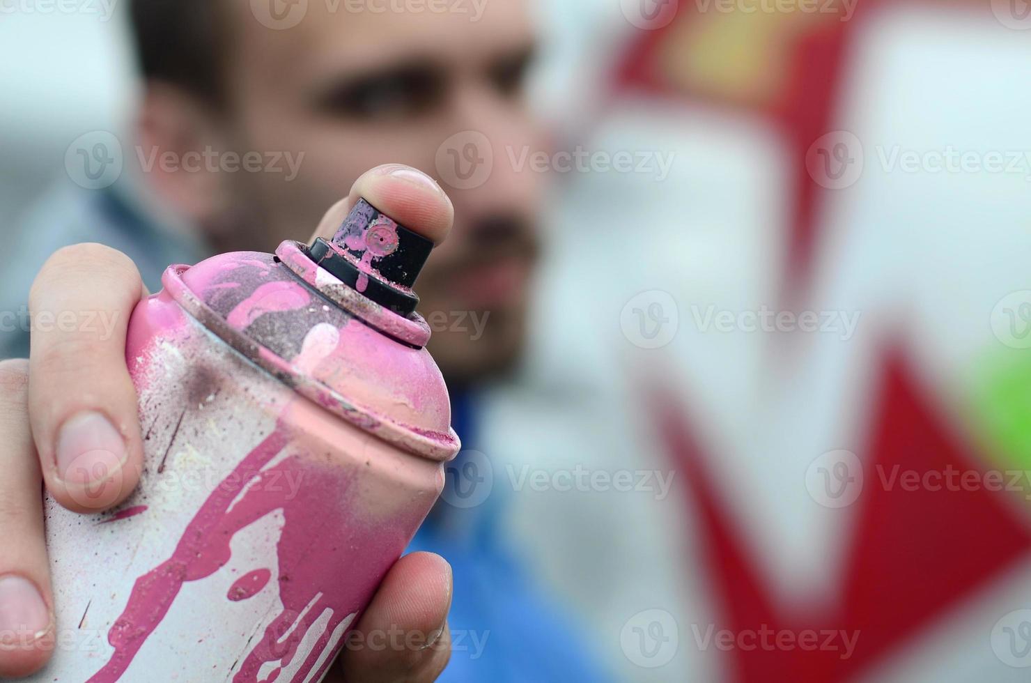 A young graffiti artist in a blue jacket is holding a can of paint in front of him against a background of colored graffiti drawing. Street art and vandalism concept photo