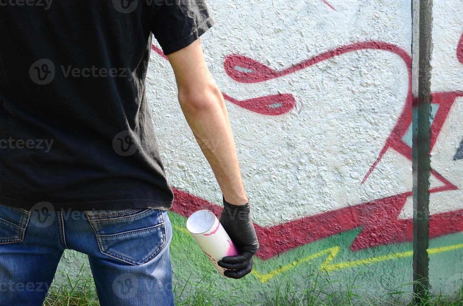 A young hooligan with a spray can stands against a concrete wall with graffiti paintings. Illegal vandalism concept. Street art photo