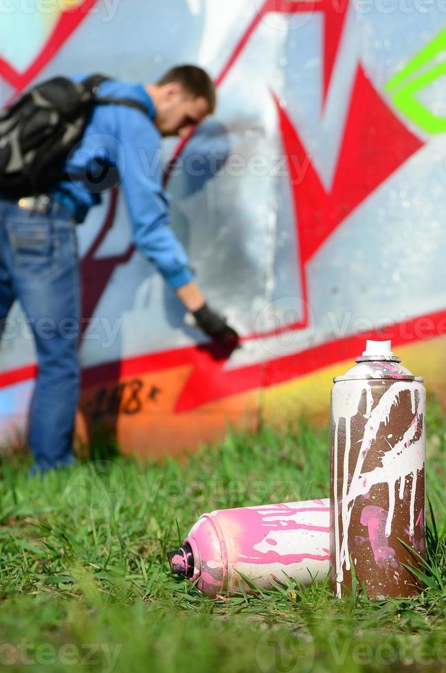 A few used paint cans against the background of the space with the wall on which the young guy draws a large graffiti drawing. Modern art of drawing walls in graffiti photo