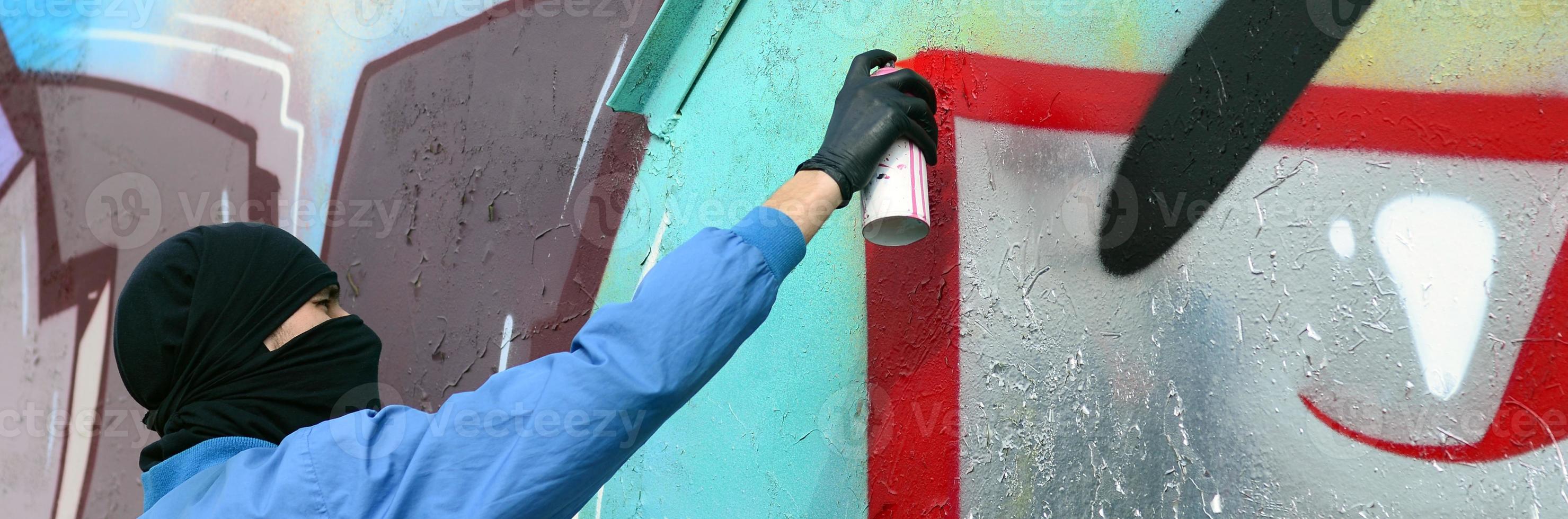 A young hooligan with a hidden face paints graffiti on a metal wall. Illegal vandalism concept photo