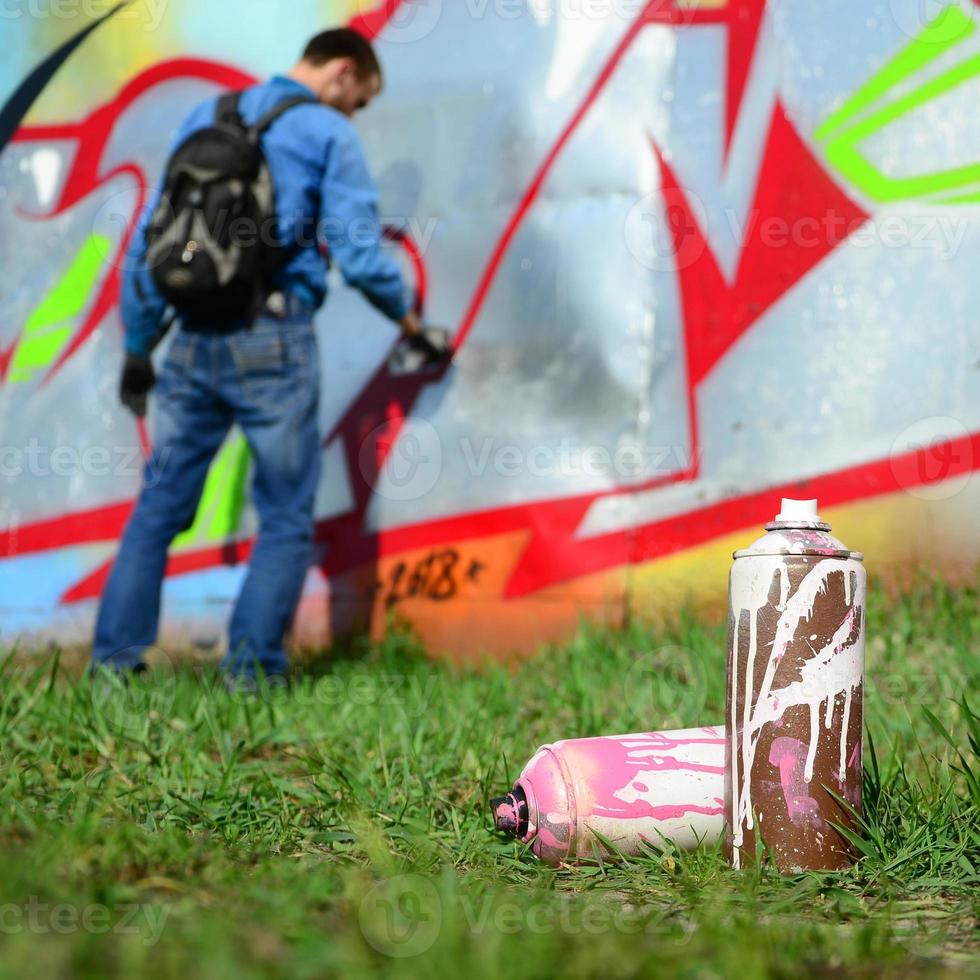 A few used paint cans against the background of the space with the wall on which the young guy draws a large graffiti drawing. Modern art of drawing walls in graffiti photo