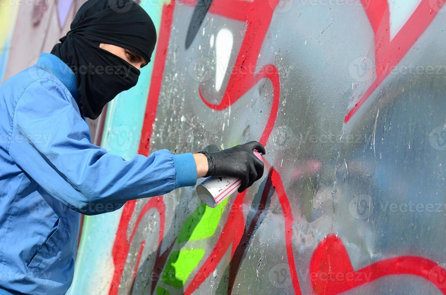 A young hooligan with a hidden face paints graffiti on a metal wall. Illegal vandalism concept photo