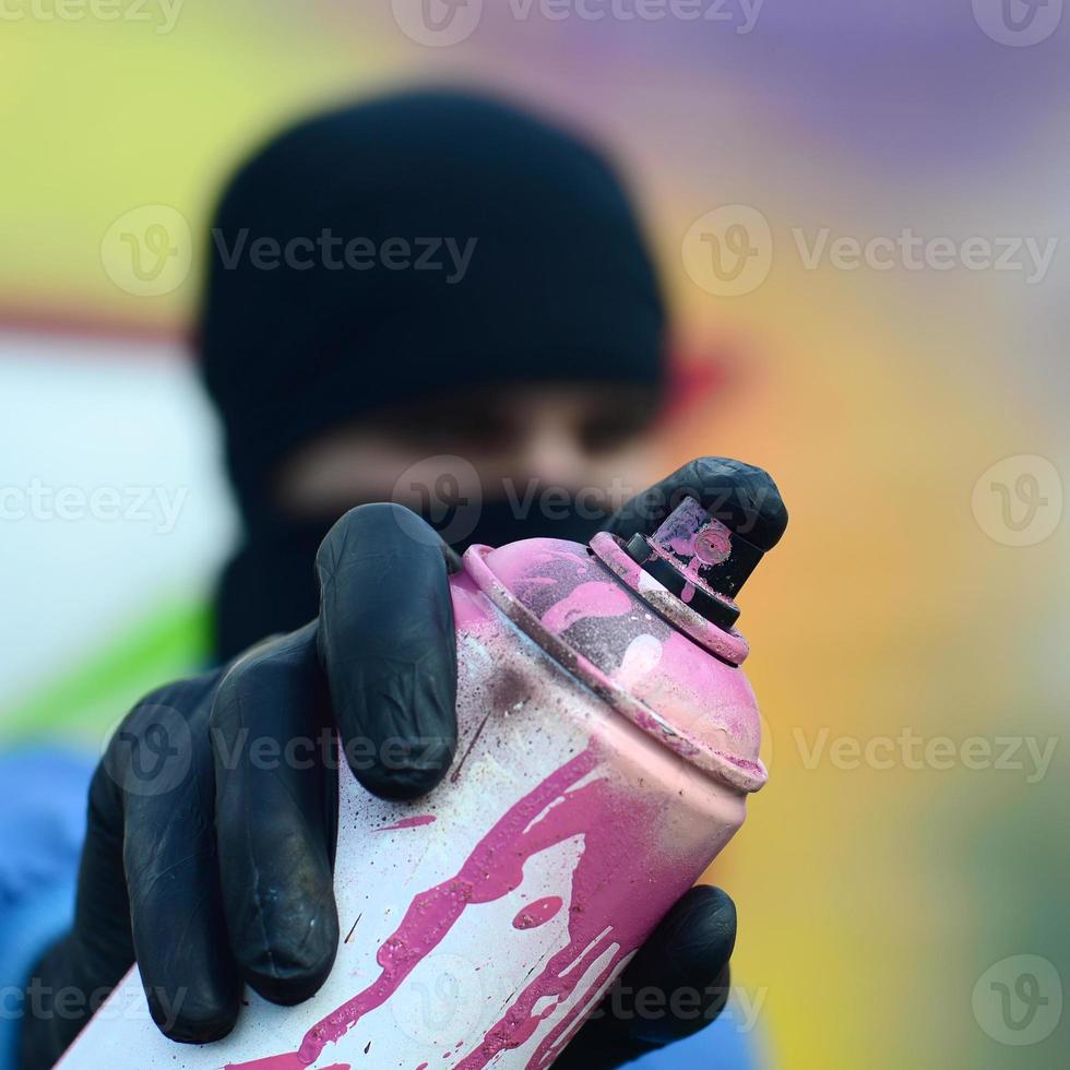 A young graffiti artist in a blue jacket and black mask is holding a can of paint in front of him against a background of colored graffiti drawing. Street art and vandalism concept photo