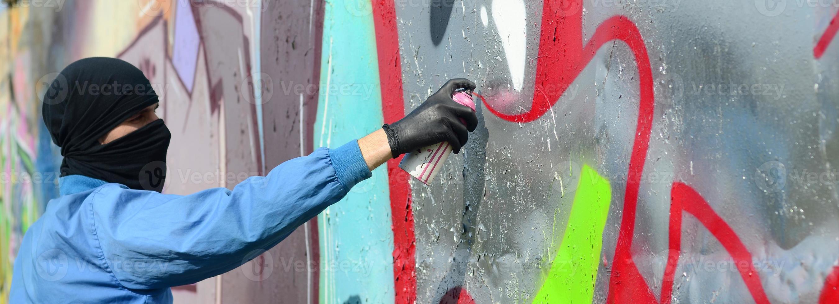 A young hooligan with a hidden face paints graffiti on a metal wall. Illegal vandalism concept photo