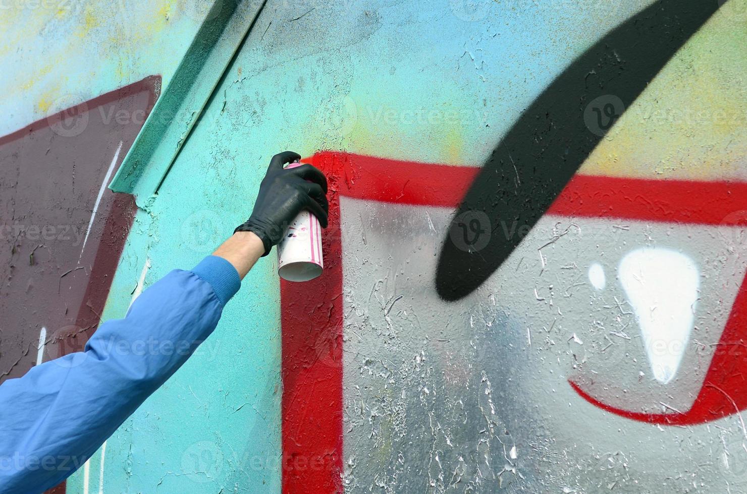A young hooligan with a hidden face paints graffiti on a metal wall. Illegal vandalism concept photo