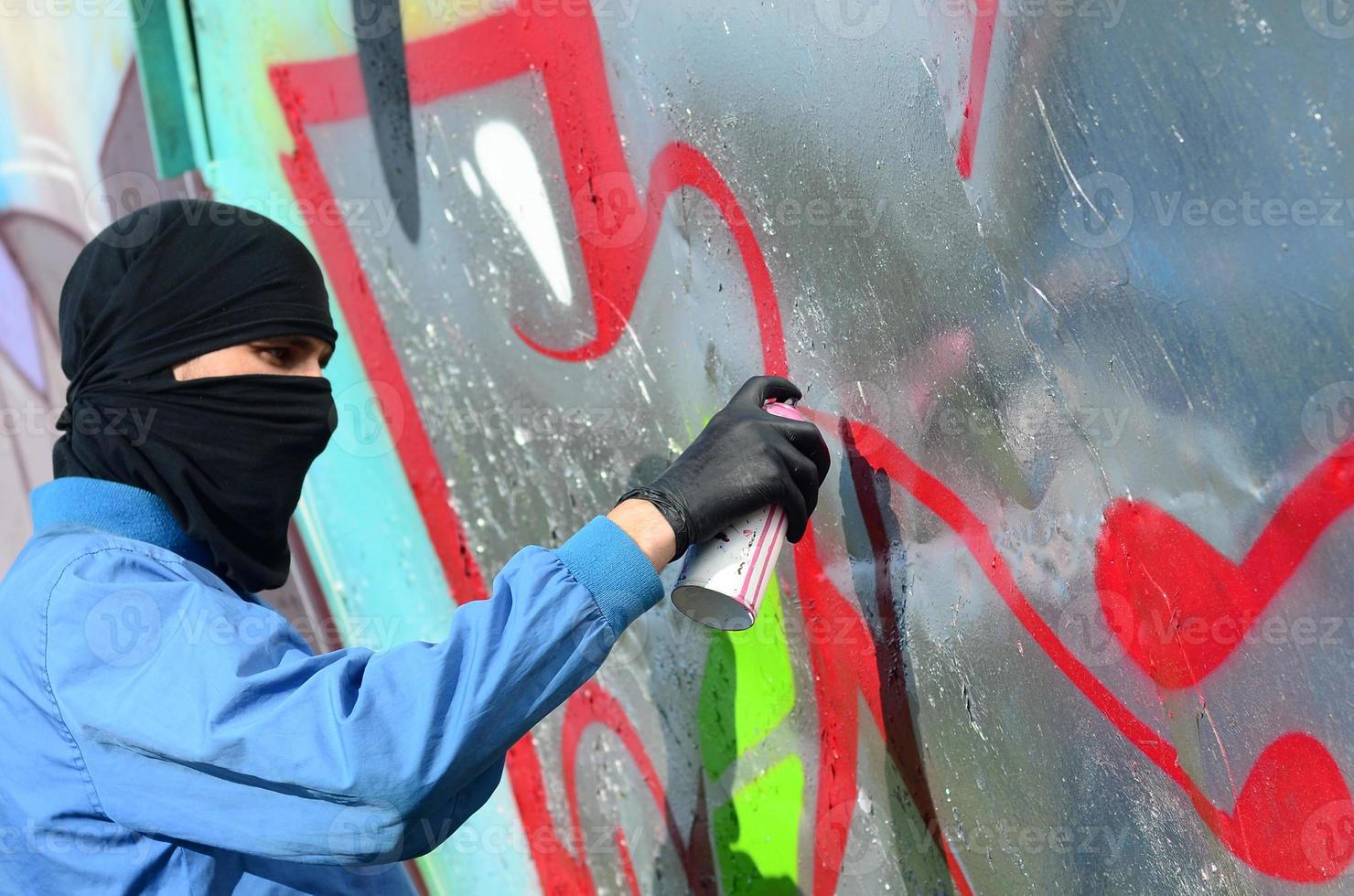 A young hooligan with a hidden face paints graffiti on a metal wall. Illegal vandalism concept photo