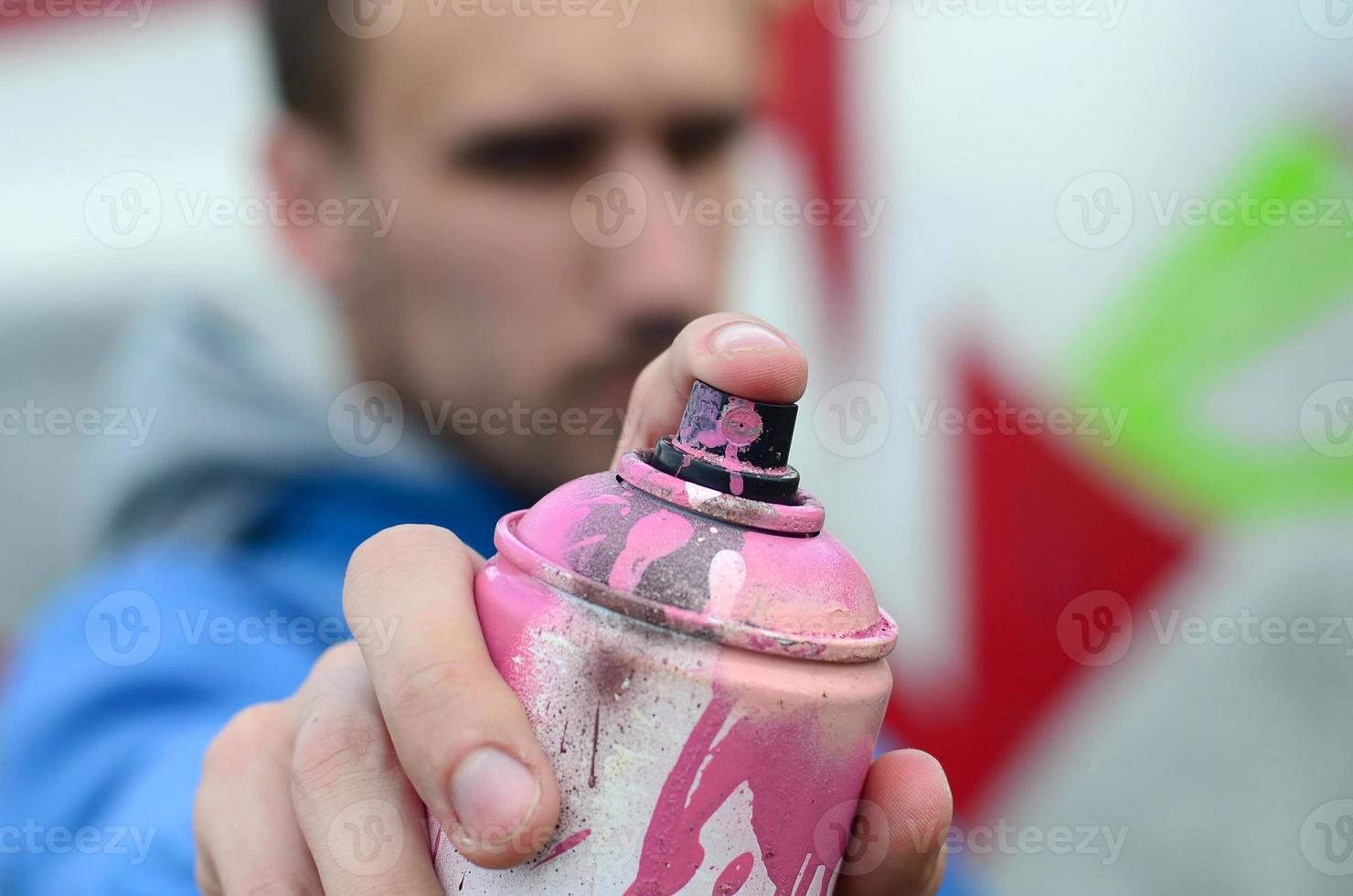 A young graffiti artist in a blue jacket is holding a can of paint in front of him against a background of colored graffiti drawing. Street art and vandalism concept photo