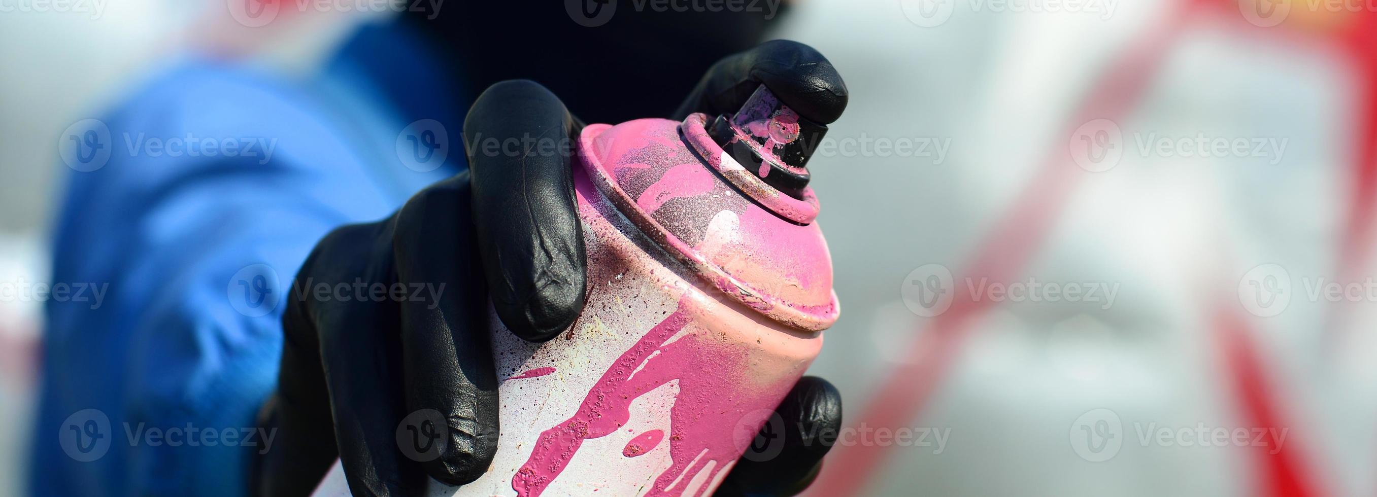 A young graffiti artist in a blue jacket and black mask is holding a can of paint in front of him against a background of colored graffiti drawing. Street art and vandalism concept photo