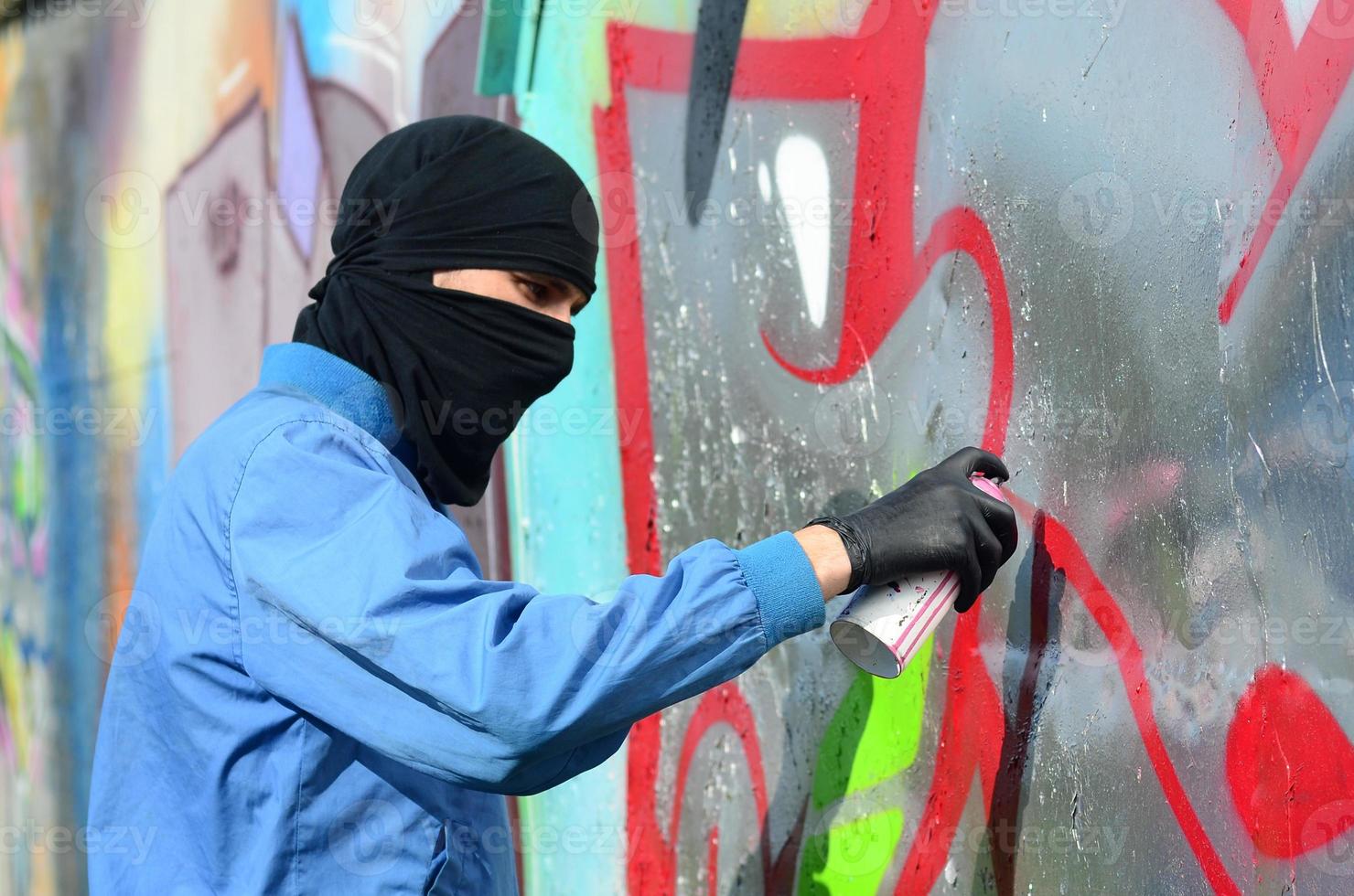 A young hooligan with a hidden face paints graffiti on a metal wall. Illegal vandalism concept photo