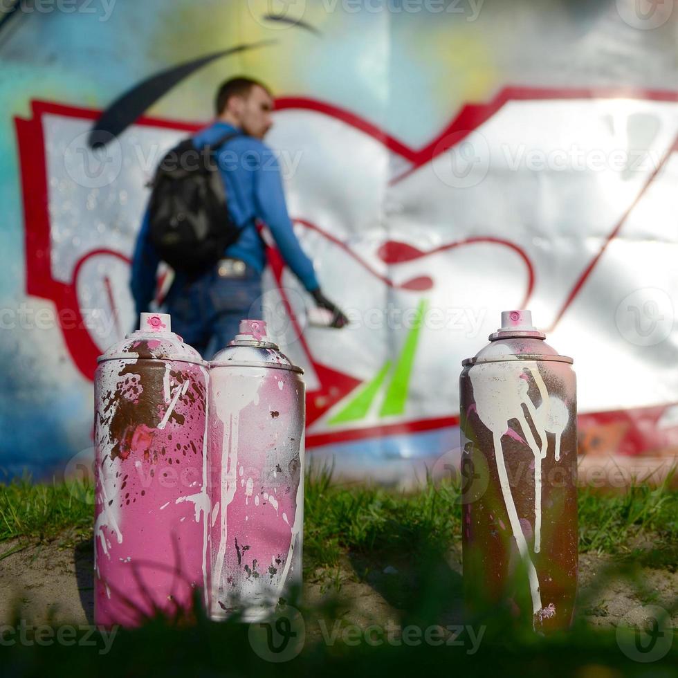 A few used paint cans against the background of the space with the wall on which the young guy draws a large graffiti drawing. Modern art of drawing walls in graffiti photo