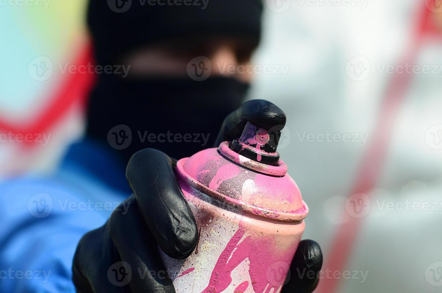A young graffiti artist in a blue jacket and black mask is holding a can of paint in front of him against a background of colored graffiti drawing. Street art and vandalism concept photo