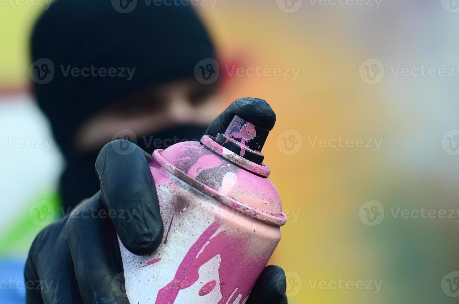 A young graffiti artist in a blue jacket and black mask is holding a can of paint in front of him against a background of colored graffiti drawing. Street art and vandalism concept photo