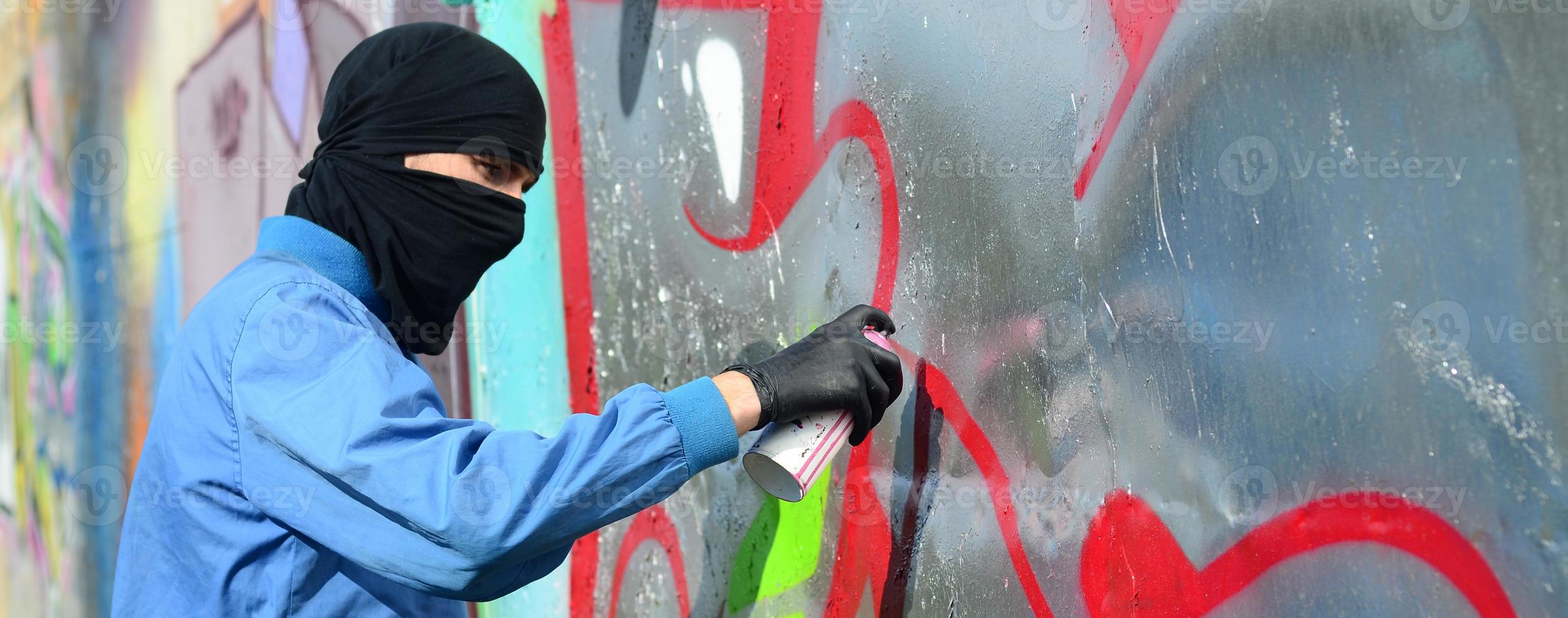 A young hooligan with a hidden face paints graffiti on a metal wall. Illegal vandalism concept photo