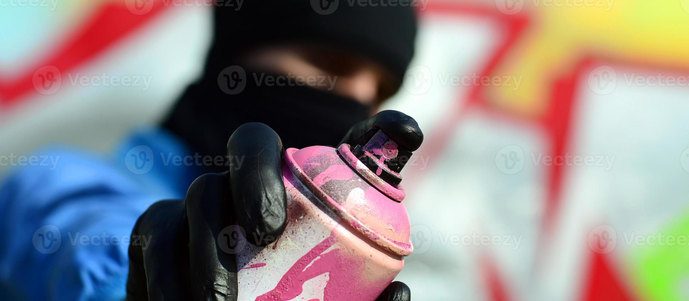 A young graffiti artist in a blue jacket and black mask is holding a can of paint in front of him against a background of colored graffiti drawing. Street art and vandalism concept photo