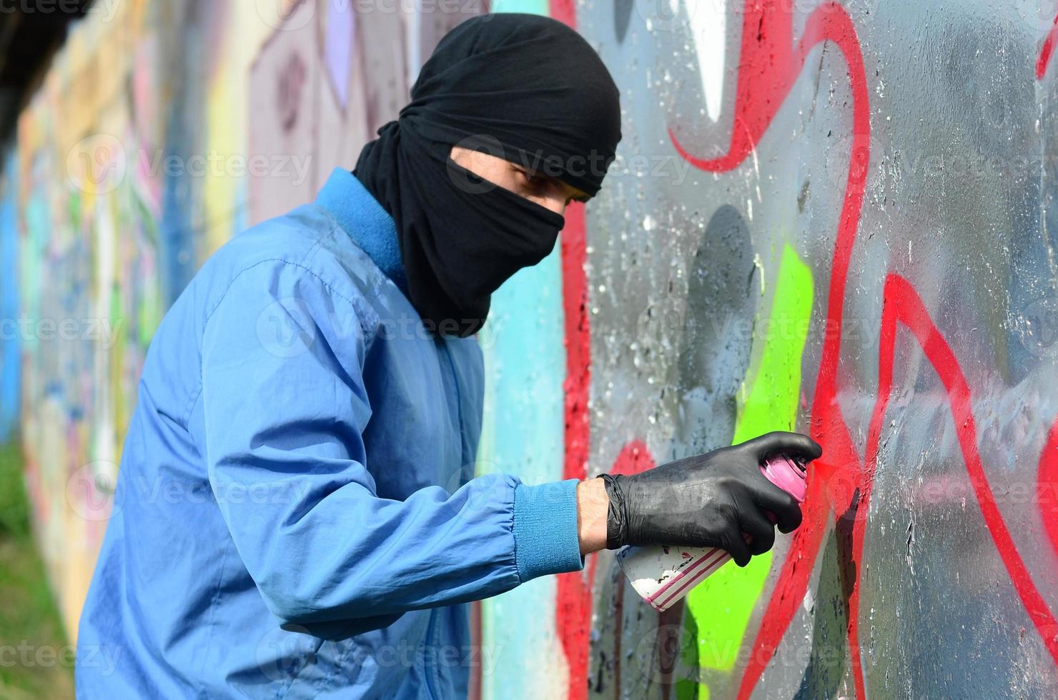 A young hooligan with a hidden face paints graffiti on a metal wall. Illegal vandalism concept photo