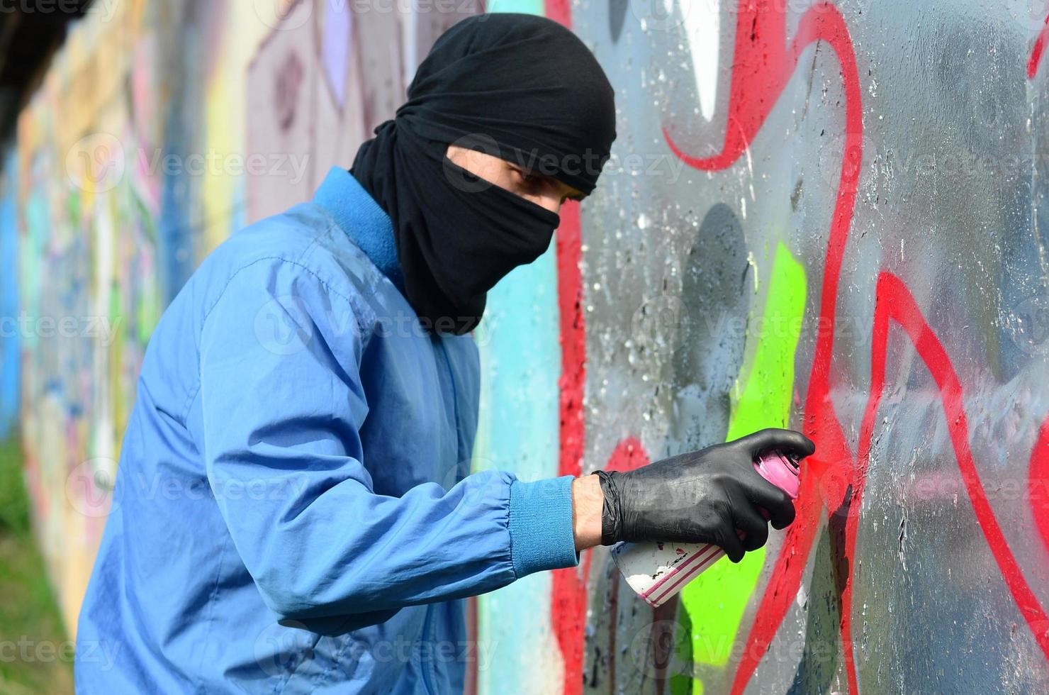 A young hooligan with a hidden face paints graffiti on a metal wall. Illegal vandalism concept photo