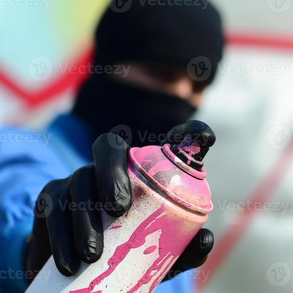 A young graffiti artist in a blue jacket and black mask is holding a can of paint in front of him against a background of colored graffiti drawing. Street art and vandalism concept photo