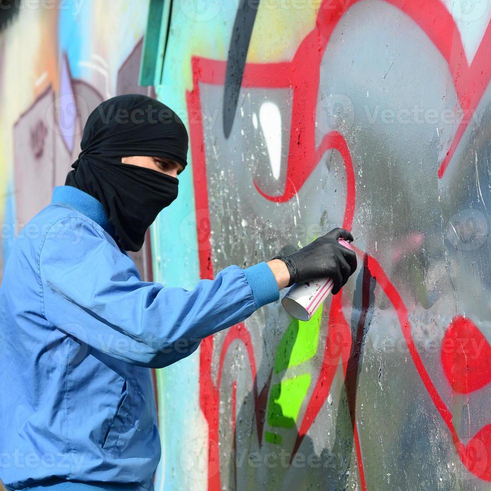 A young hooligan with a hidden face paints graffiti on a metal wall. Illegal vandalism concept photo