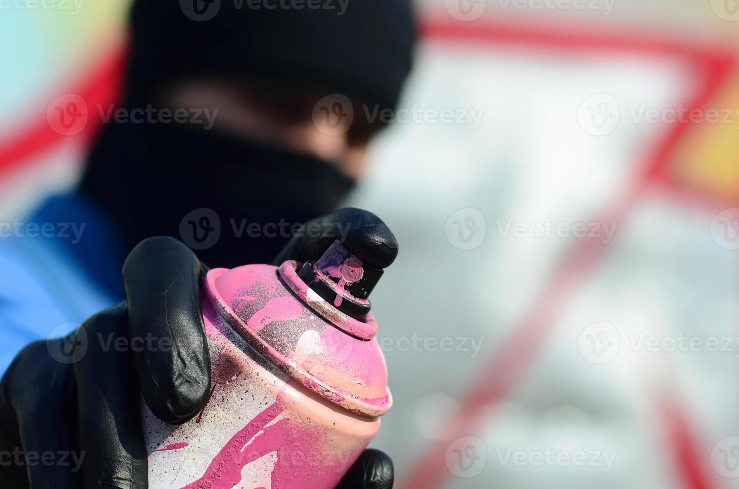 A young graffiti artist in a blue jacket and black mask is holding a can of paint in front of him against a background of colored graffiti drawing. Street art and vandalism concept photo