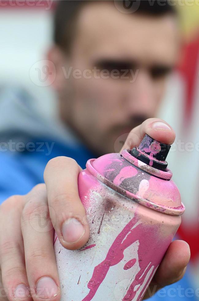 A young graffiti artist in a blue jacket is holding a can of paint in front of him against a background of colored graffiti drawing. Street art and vandalism concept photo