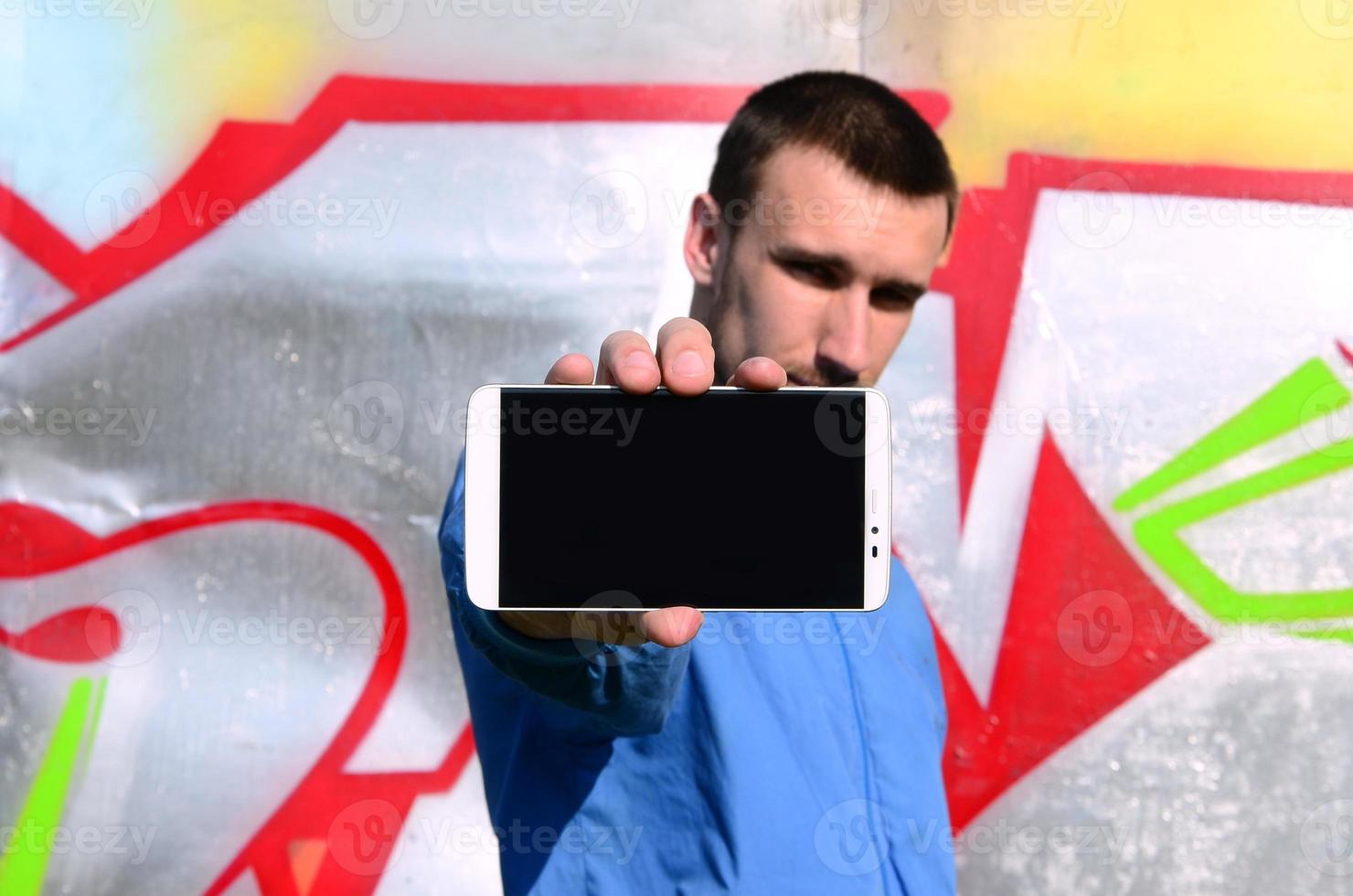 The graffiti artist demonstrates a smartphone with an empty black screen against the background of a colorful painted wall. Street art concept photo