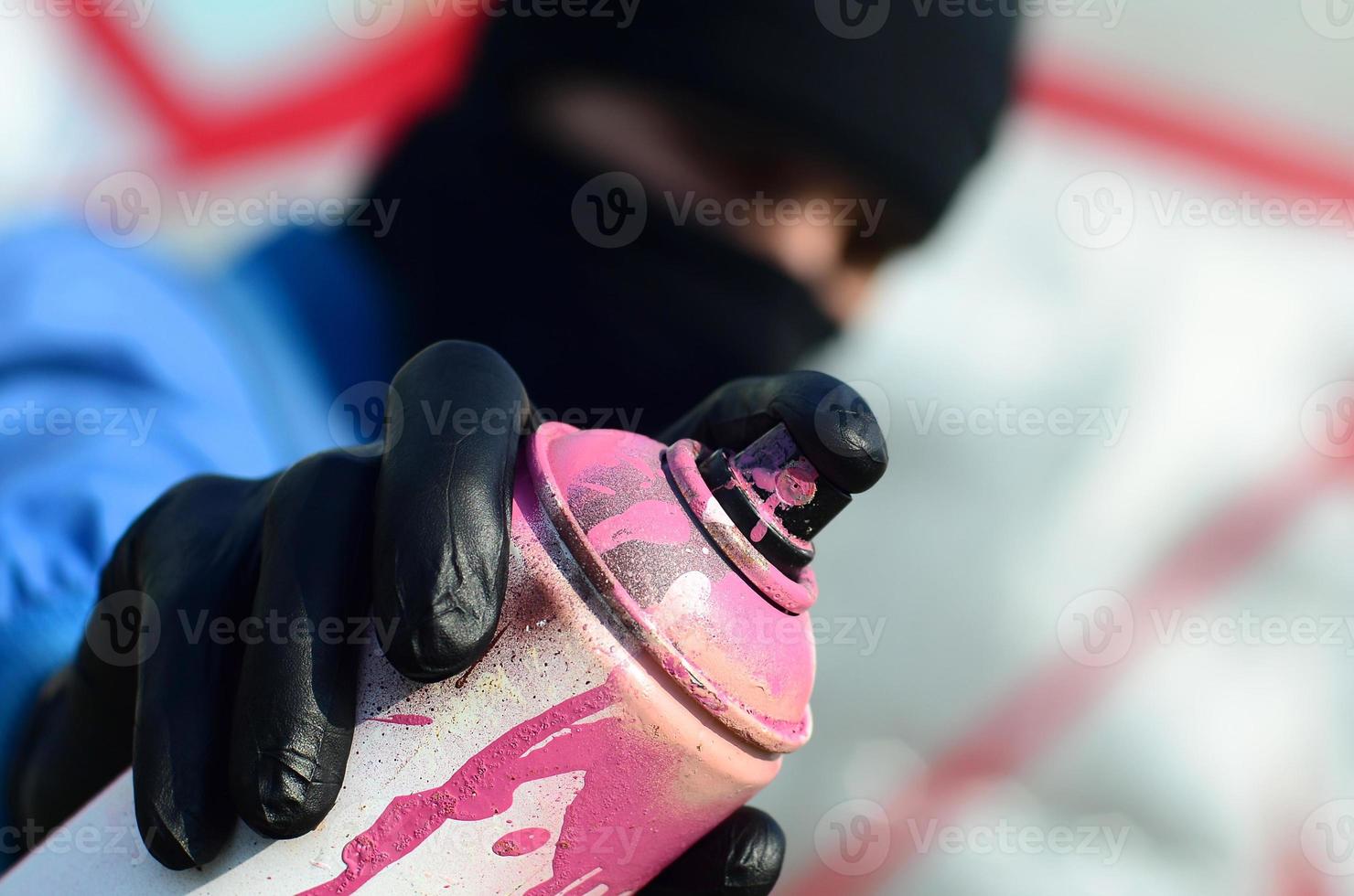 A young graffiti artist in a blue jacket and black mask is holding a can of paint in front of him against a background of colored graffiti drawing. Street art and vandalism concept photo