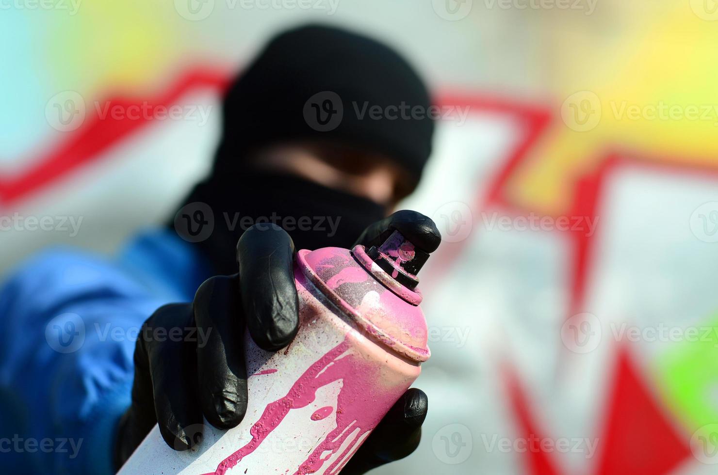 un joven artista de graffiti con una chaqueta azul y una máscara negra sostiene una lata de pintura frente a él contra un fondo de dibujo de graffiti de colores. concepto de arte callejero y vandalismo foto