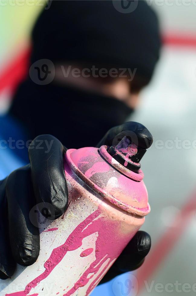 A young graffiti artist in a blue jacket and black mask is holding a can of paint in front of him against a background of colored graffiti drawing. Street art and vandalism concept photo