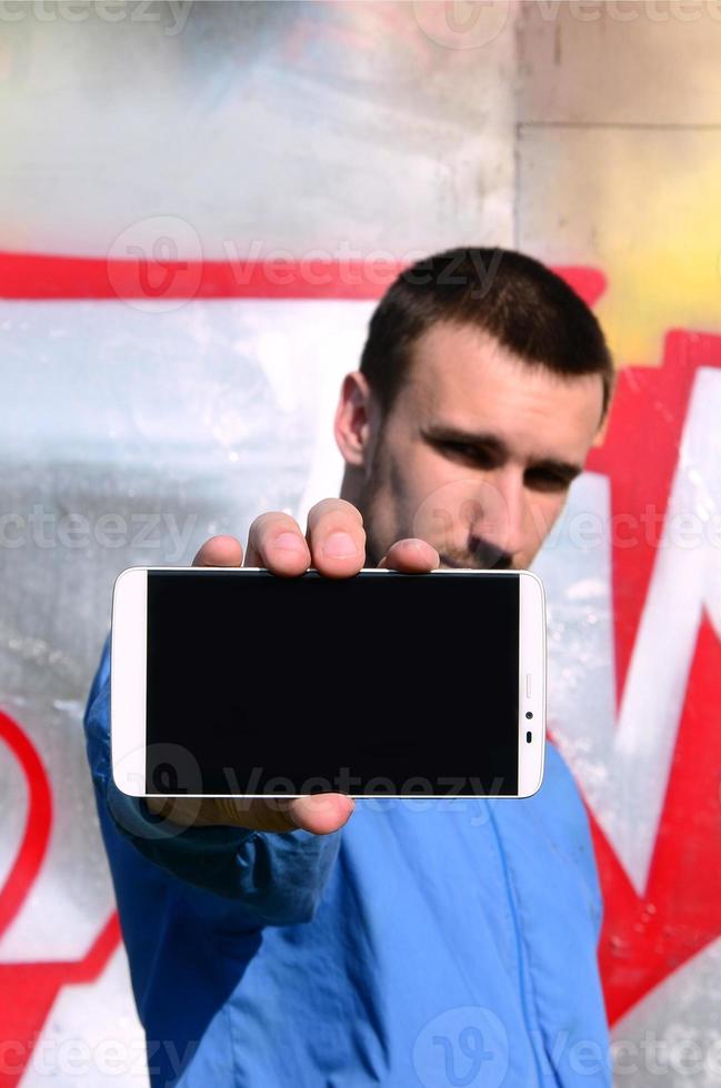 The graffiti artist demonstrates a smartphone with an empty black screen against the background of a colorful painted wall. Street art concept photo