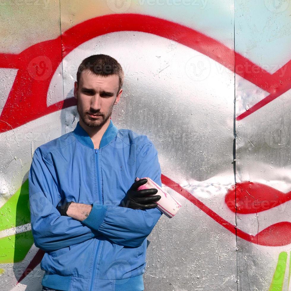 el artista de graffiti con lata de aerosol posa contra el fondo de una pared pintada de colores. concepto de arte callejero foto