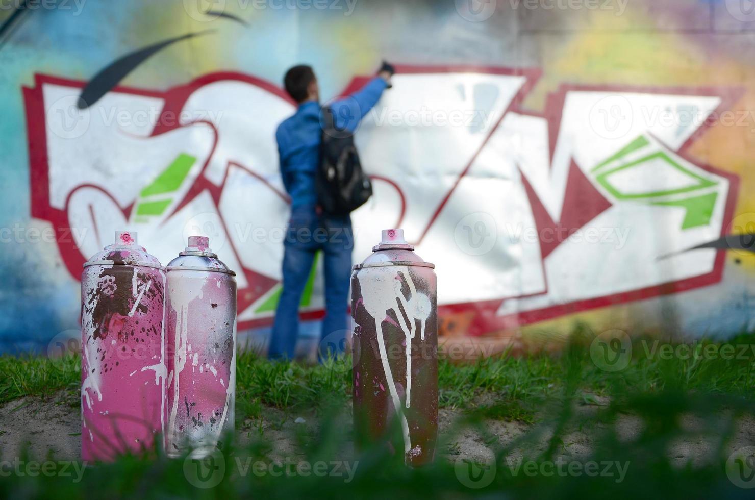 A few used paint cans against the background of the space with the wall on which the young guy draws a large graffiti drawing. Modern art of drawing walls in graffiti photo