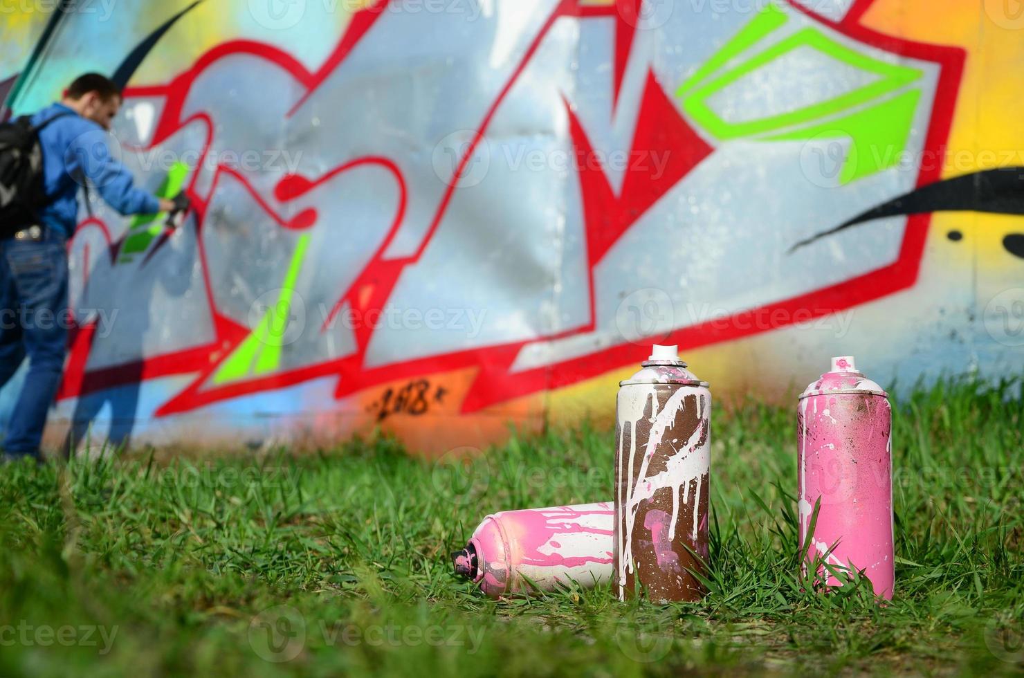 A few used paint cans against the background of the space with the wall on which the young guy draws a large graffiti drawing. Modern art of drawing walls in graffiti photo