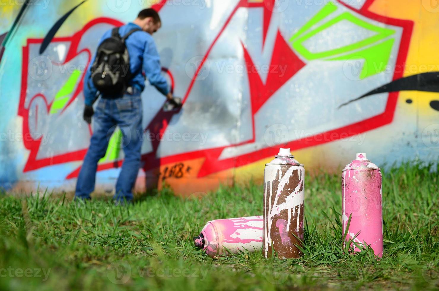 A few used paint cans against the background of the space with the wall on which the young guy draws a large graffiti drawing. Modern art of drawing walls in graffiti photo