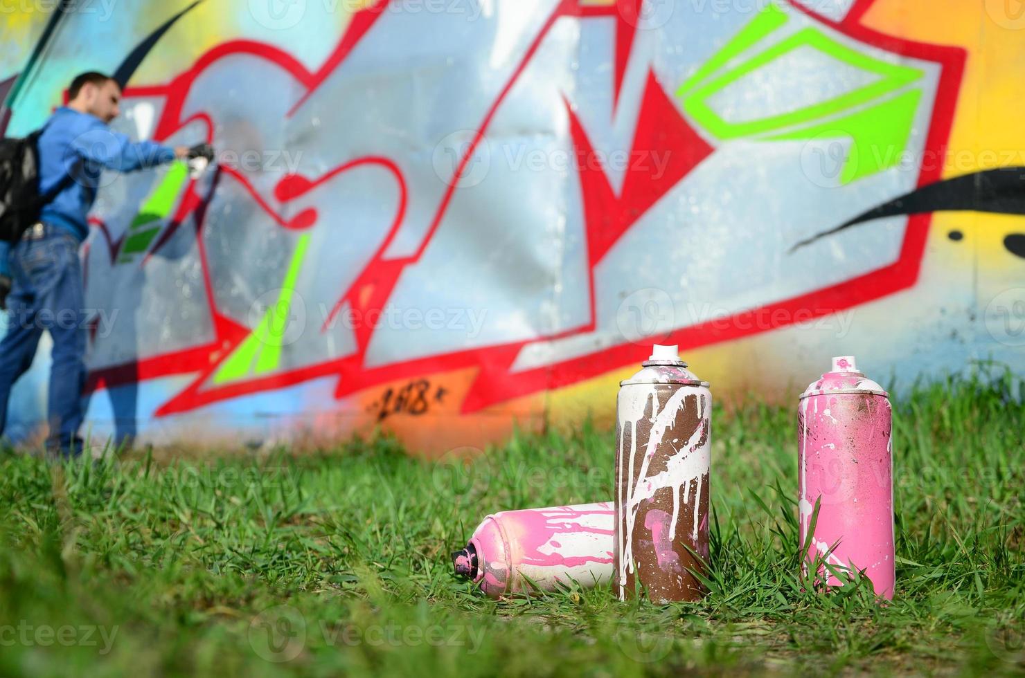 A few used paint cans against the background of the space with the wall on which the young guy draws a large graffiti drawing. Modern art of drawing walls in graffiti photo